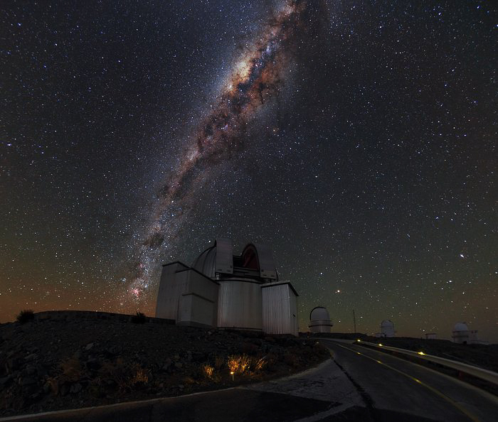 High up in the Chilean Atacama Desert, pioneering feats of human engineering collide with the majestic beauty of the natural world. This image shows ESO’s La Silla Observatory, where domes housing some of the most advanced astronomical instruments in the world sit beneath a sky shimmering with stars. All of these stars belong to our home galaxy, the Milky Way. The Milky Way contains billions of stars, arranged in two strikingly different structures. The roughly spherical halo component, consisting mainly of older stars, appears in this image as the background of stars scattered across the sky. The second component is a thin disc made up of younger stars, gas and dust. We see this as a dense, bright, and visually stunning band running almost vertically across the sky. Pockets of dust block out the light from stars behind, giving the band a mottled appearance. The bright concentration in the band of stars, located toward the top centre of this image, is the central region of the Milky Way. Here, astronomers have measured stars moving very much faster than anywhere else in our galaxy. This is taken as evidence for a supermassive black hole, some four million times the mass of the Sun, at the very centre of our galaxy. The black hole cannot be observed directly, but its presence can be inferred from the effect its enormous gravity has on the motions of these nearby stars.